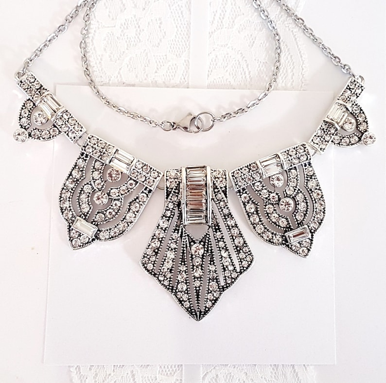 Puede incluir: Un collar de plata con un dise&ntilde;o geom&eacute;trico que presenta un colgante central en forma de diamante y dos colgantes m&aacute;s peque&ntilde;os a cada lado. El collar est&aacute; adornado con diamantes de imitaci&oacute;n transparentes.