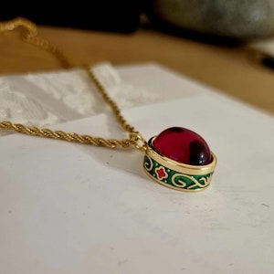 May include: A gold chain necklace with a red oval pendant. The pendant has a green and gold filigree design.