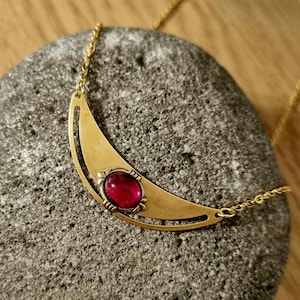 May include: A gold crescent moon necklace with a red gemstone pendant. The necklace is on a gray stone background.
