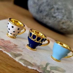 Könnte beinhalten: Drei Miniatur-Teetassen mit goldenen Verzierungen. Eine ist weiß mit schwarzen Punkten, eine ist blau mit goldenen Akzenten und eine ist hellblau.