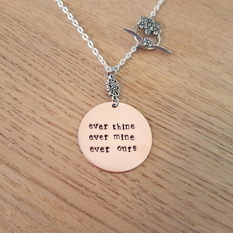 Pode incluir: Colar de prata com um pingente circular cor de cobre. O pingente tem as palavras "ever thine ever mine ever ours" gravadas. O colar tem um fecho de altern&acirc;ncia floral. Fundo de madeira.