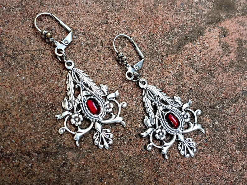 May include: A pair of silver earrings with red gemstone accents. The earrings feature a delicate floral design with intricate details.