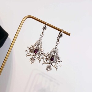 May include: Silver dangle earrings with intricate floral designs and a dark purple gemstone in the center of each earring.