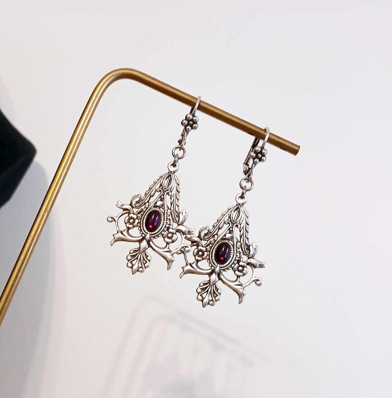 May include: Silver dangle earrings with intricate floral designs and a dark purple gemstone in the center of each earring.