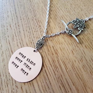 Pode incluir: Colar de prata com um pingente oval cor de cobre. O pingente tem as palavras "ever thine, ever mine, ever ours". O colar tem um fecho floral e detalhes decorativos.