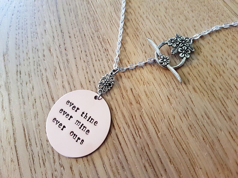 Pode incluir: Colar de prata com um pingente oval cor de cobre. O pingente tem as palavras "ever thine, ever mine, ever ours". O colar tem um fecho floral e detalhes decorativos.