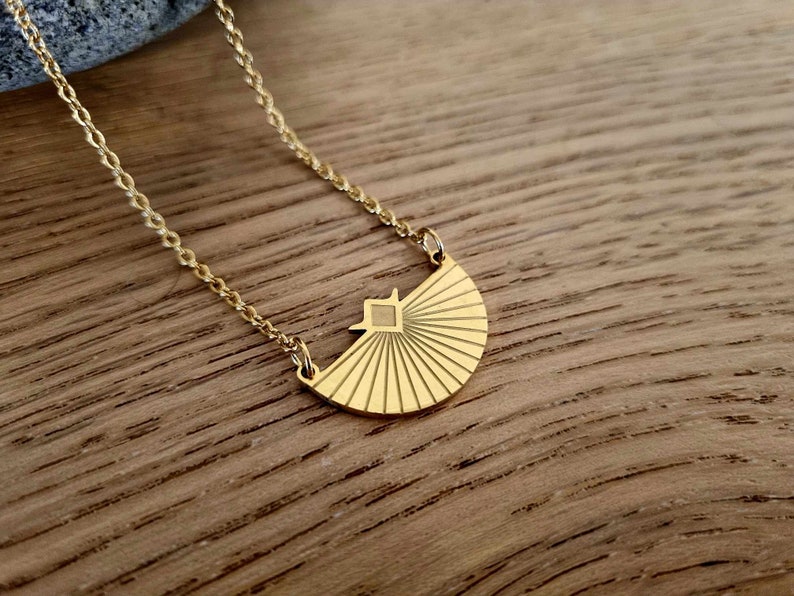 May include: A gold necklace with a geometric sunburst pendant. The pendant is made of metal and has a textured surface.