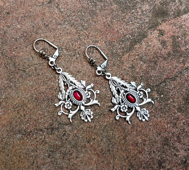 May include: A pair of silver earrings with red gemstone accents. The earrings feature a delicate floral design.