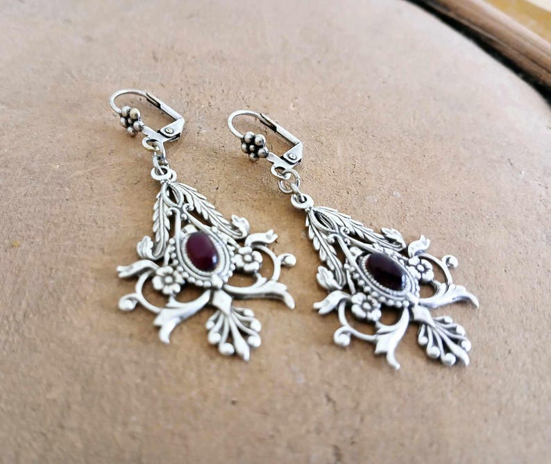 May include: A pair of silver dangle earrings with intricate floral designs and a dark red gemstone in the center of each earring.