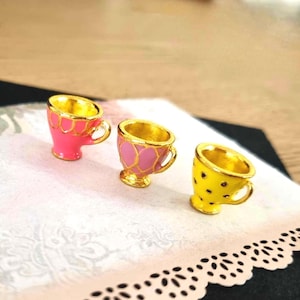 Könnte beinhalten: Drei Miniatur-Dekotassen in Rosa, Gelb und Pink mit goldenen Akzenten. Die Tassen haben ein goldenes Innenleben und Henkel. Die rosa Tasse hat ein Muschelmuster, die zweite rosa Tasse ein Rautenmuster und die gelbe Tasse schwarze Tupfen.
