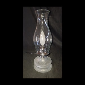 May include: A clear glass electric lamp with a frosted base and a clear glass chimney. The base has a textured design, and the lamp has a silver-colored metal accent. The light bulb is shaped like a flame.