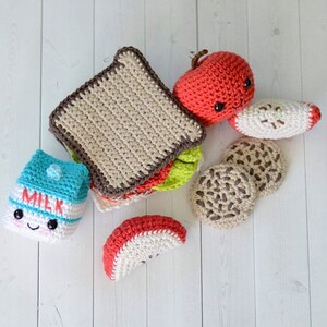 May include: A collection of crocheted play food items including a sandwich with brown crust, a red apple, two apple slices, two chocolate chip cookies, and a small blue and white milk carton with the word MILK embroidered in red on the front.