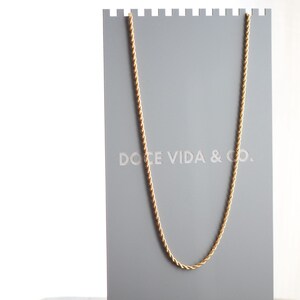 May include: A gold rope chain necklace displayed on a gray stand with the text "DOCE VIDA & CO."