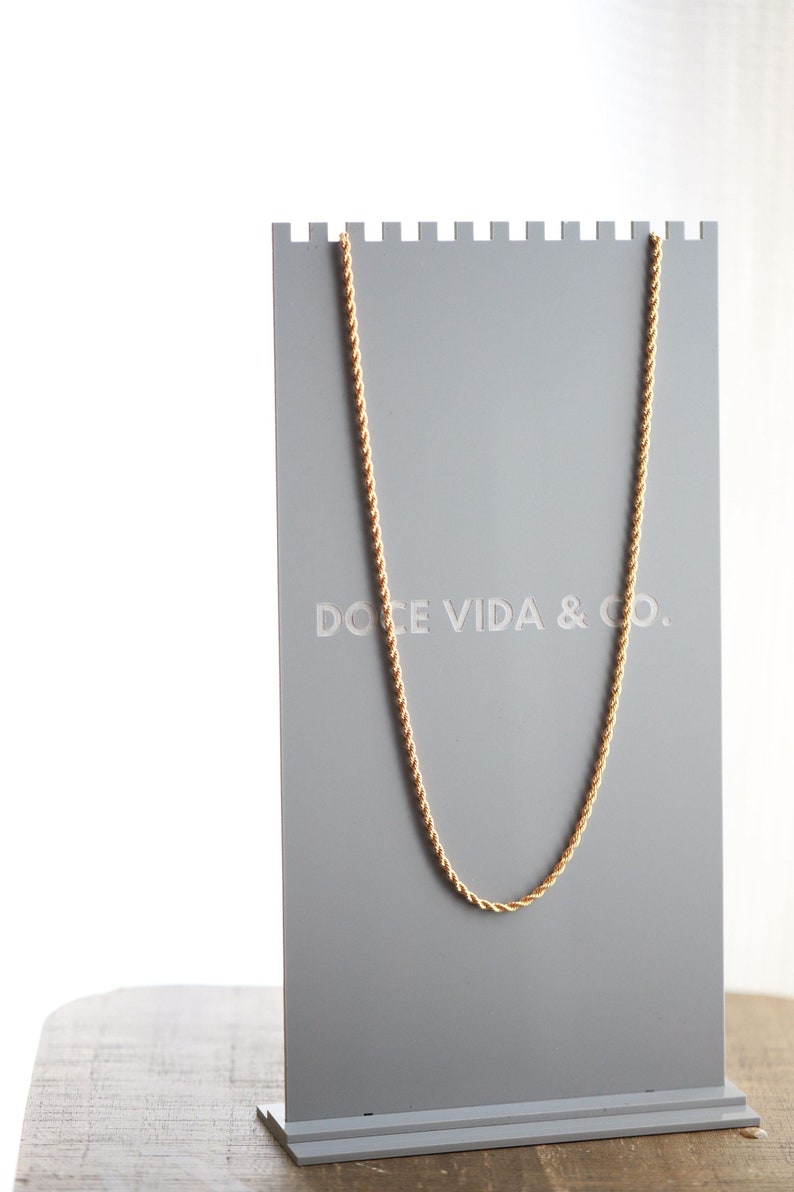 May include: A gold rope chain necklace displayed on a gray stand with the text "DOCE VIDA & CO."