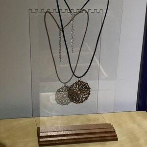 May include: A wooden pendant necklace with a geometric design hangs on a black cord from a clear acrylic display stand with a wooden base.