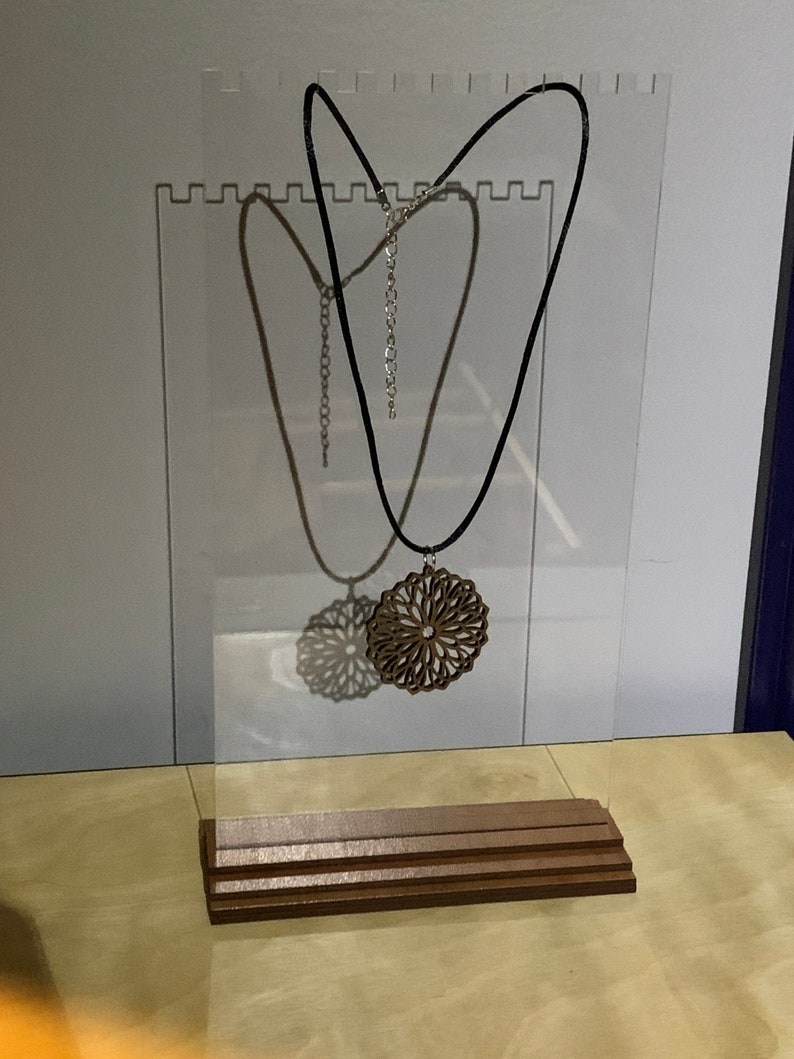May include: A wooden pendant necklace with a geometric design hangs on a black cord from a clear acrylic display stand with a wooden base.