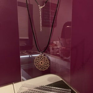 May include: A wooden pendant necklace with a delicate, intricate design hangs from a black cord on a clear acrylic display stand.