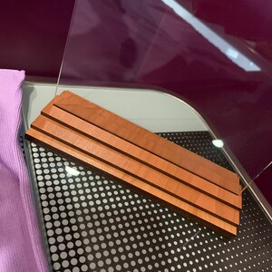 May include: A clear acrylic display stand with a wooden base. The base is made of three layers of wood, each with a different grain pattern. The stand is sitting on a black surface with white polka dots.