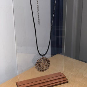 May include: A wooden pendant necklace with a detailed mandala design hangs on a clear acrylic display stand with a wooden base.