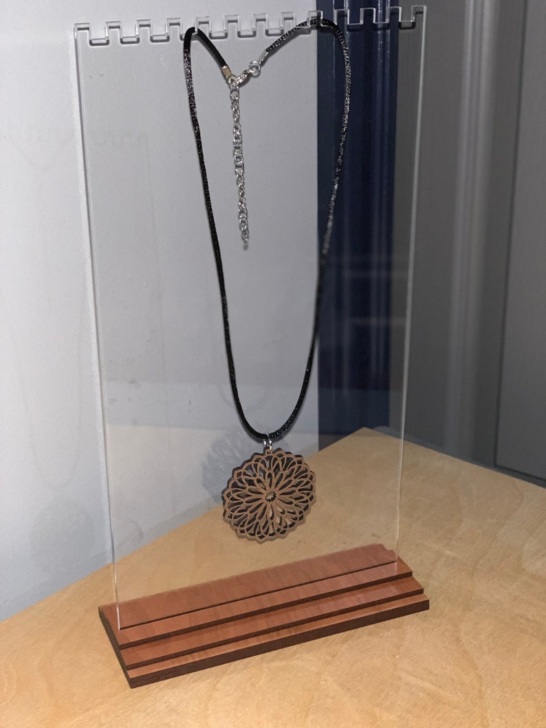 May include: A wooden pendant necklace with a detailed mandala design hangs on a clear acrylic display stand with a wooden base.