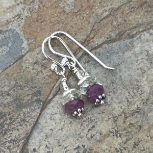May include: A pair of silver dangle earrings. Each earring features a light green stone, a deep purple stone, and small silver accents. The earrings are displayed on a textured, neutral-toned surface.