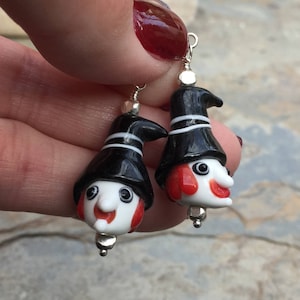 May include: A pair of dangle earrings featuring black and white glass witch figures with red noses and white eyes. The figures are wearing black hats with white stripes.