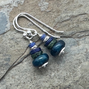 Puede incluir: Un par de pendientes hechos a mano con ganchos plateados. Cada pendiente presenta una pila de cuentas de piedras preciosas azules y verdes, incluyendo formas redondas y cuadradas. Los pendientes se exhiben sobre una superficie gris texturizada.