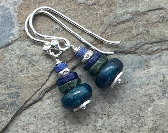 Op de afbeelding: Een paar handgemaakte oorbellen met zilverkleurige haken. Elke oorbel heeft een stapel blauwe en groene edelsteen kralen, waaronder ronde en vierkante vormen. De oorbellen worden gepresenteerd op een getextureerd, grijs oppervlak.