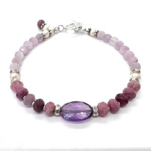 Puede incluir: Una pulsera delicada con una variedad de piedras preciosas moradas y rosas. Incluye una amatista ovalada, cuentas redondas y pequeñas perlas, todo ensartado con un cierre plateado. La paleta de colores es suave y femenina.