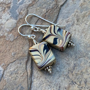 Puede incluir: Un par de pendientes de plata con cuentas cuadradas de vidrio marmoleado en negro, blanco y marrón. Las cuentas tienen un patrón de remolino.