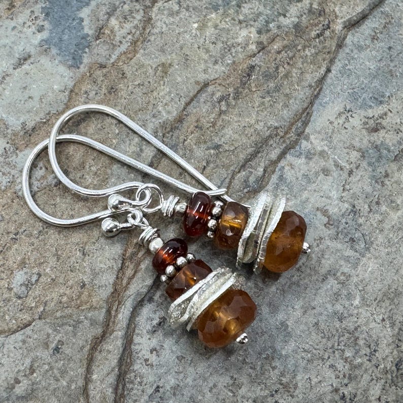 Op de afbeelding: Een paar handgemaakte oorbellen met zilveren haken en ingewikkeld kralenwerk. De oorbellen tonen gefacetteerde, amberkleurige edelstenen en kleinere, donkerrode kralen, geaccentueerd met zilveren ringen. Het ontwerp is geplaatst tegen een getextureerde, grijze stenen achtergrond.