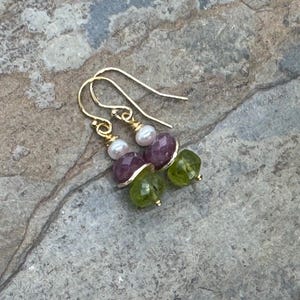 May include: Gold-toned earrings with a drop design. Each earring features a small white pearl, a faceted purple bead, and a faceted green bead. The earrings are displayed on a gray stone surface.