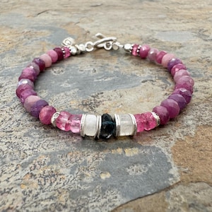Könnte beinhalten: Armband mit rosa und lila Edelsteinen. Das Armband hat einen Farbverlauf, mit facettierten und abgerundeten Perlen, silberfarbenen Akzenten und einem Verschluss. In der Mitte des Armbands befinden sich weiße und dunkelblaue Steine.