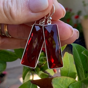 Puede incluir: Un par de pendientes de plata con gotas rectangulares de piedra preciosa roja. Los pendientes tienen un diseño facetado y son de color rojo intenso.
