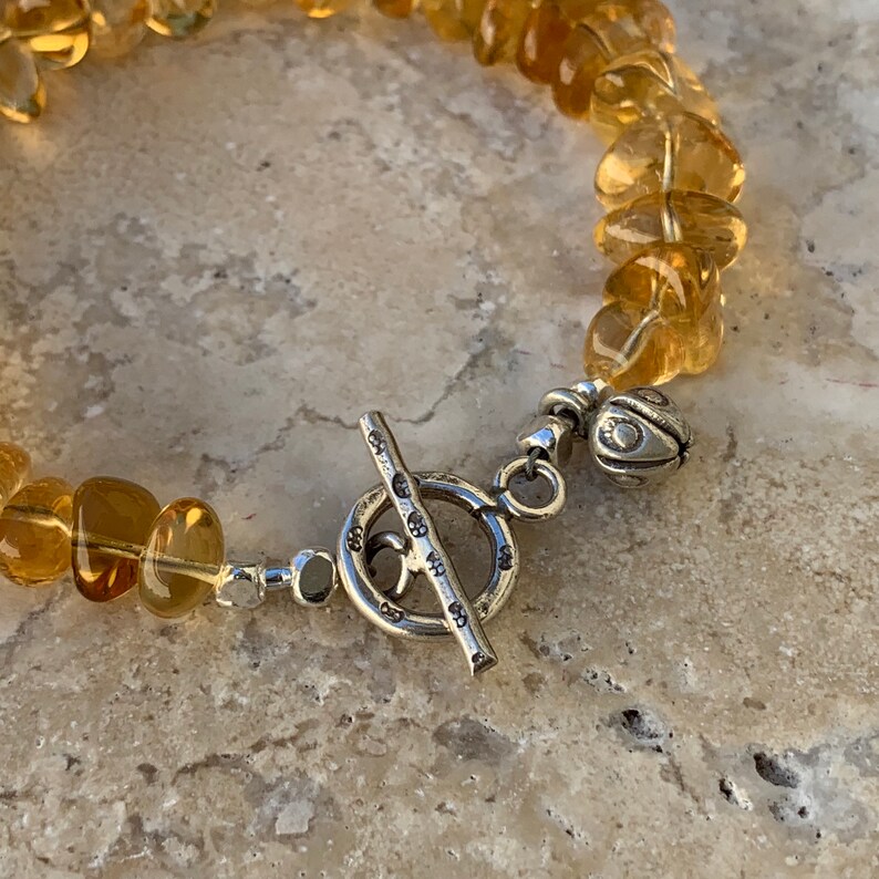 May include: A bracelet made of small, faceted, amber-colored stones with a silver toggle clasp. The clasp has a decorative design with small circles.