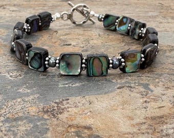 May include: A bracelet featuring square-shaped abalone shell beads with iridescent green, blue, and purple tones. The beads are separated by small metallic silver-colored spheres and dark grey beads, finished with a silver-toned metal toggle clasp.
