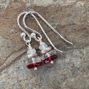 May include: A pair of silver drop earrings featuring stacked beads. Each earring includes a translucent white bead, a textured silver spacer, a clear faceted crystal bead, and a deep red faceted crystal bead at the base, hanging from silver hooks.