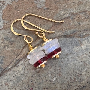 May include: A pair of gold-toned hook earrings featuring stacked faceted beads. Each earring consists of two translucent white moonstone-like discs layered above two deep red faceted gemstone discs, secured with gold wire and small beads.