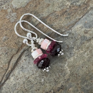 May include: A pair of dangle earrings with silver hooks. Each earring features a cluster of beads in shades of pink and deep red, with a faceted, dark red bead at the bottom. The earrings are set against a textured, gray background.