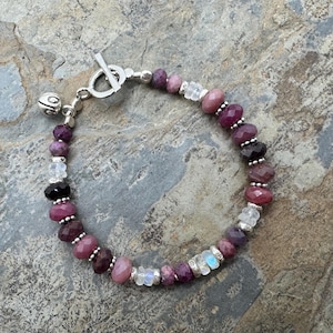 Peut inclure: Un bracelet de perles avec des pierres précieuses roses, violettes et transparentes. Le bracelet est orné d'un fermoir à bascule et d'un petit grelot argenté. Les perles à facettes sont enfilées avec de petites perles argentées.