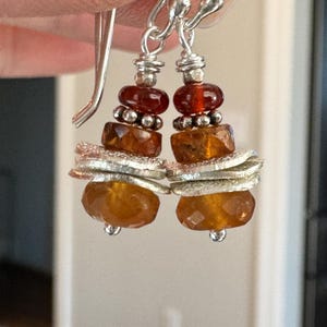 Op de afbeelding: Hangende oorbellen met zilveren haken. De oorbellen hebben een gelaagd ontwerp van oranje en zilveren kralen, met facetgeslepen en ronde kralen. De bovenste kralen zijn dieprood.