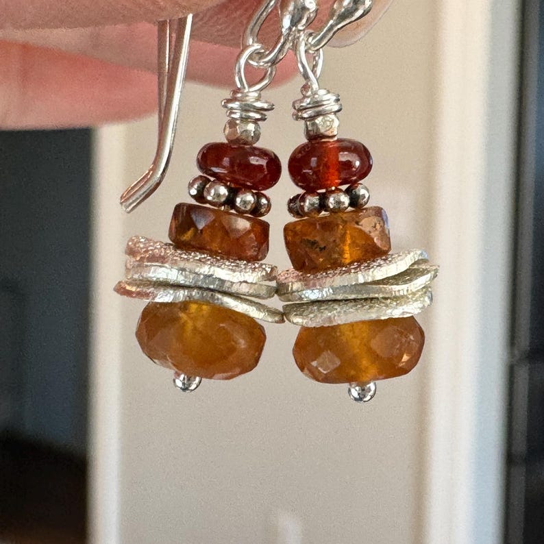 Op de afbeelding: Hangende oorbellen met zilveren haken. De oorbellen hebben een gelaagd ontwerp van oranje en zilveren kralen, met facetgeslepen en ronde kralen. De bovenste kralen zijn dieprood.