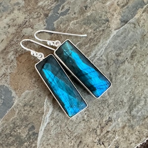 Puede incluir: Un par de pendientes rectangulares con piedras de labradorita pulidas engastadas en marcos de metal plateado. Cada piedra muestra un destello azul iridiscente vibrante en su superficie, suspendidas de sencillos ganchos plateados.