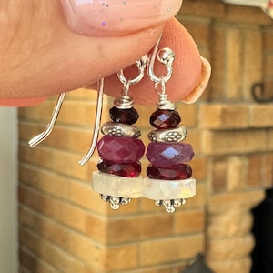May include: Dangling earrings with a silver hook and a layered design. Each earring features a dark red gemstone at the top, followed by silver beads, a faceted pink gemstone, and a white gemstone at the bottom. The earrings are held up by a silver hook.