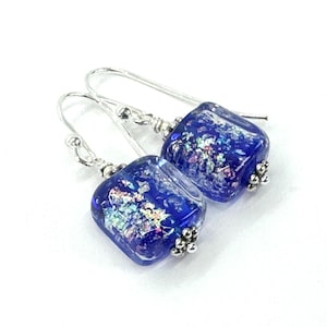 May include: A pair of square, dichroic glass earrings in a deep blue hue. The glass has iridescent flecks that catch the light. The earrings have silver-tone ear wires and small, decorative silver-tone beads.