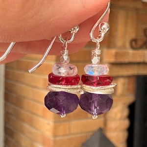 May include: A pair of dangle earrings featuring stacked faceted beads in purple, red, and iridescent white, separated by thin silver-toned metal spacers. The earrings hang from silver-toned metal hooks and display a multi-layered design.