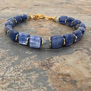 Puede incluir: Una pulsera con cuentas rectangulares y lisas de cianita azul separadas por pequeños espaciadores de metal dorado. La pieza incluye un cierre de palanca de metal dorado y una cuenta facetada y translúcida cerca del mecanismo de cierre.