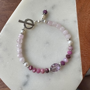 Könnte beinhalten: Ein handgefertigtes Armband mit Knebelverschluss, das eine Mischung aus rosa und lila Edelsteinen zeigt. Es enthält facettierten Rosenquarz, Perlen und Amethystperlen mit silberfarbenen Akzenten. Die Farbpalette ist weich und feminin.