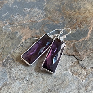 May include: A pair of rectangular amethyst earrings with a faceted design. The earrings have a silver-colored metal border and a hook closure. The earrings are displayed on a gray stone surface.