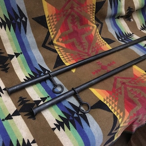 May include: Two black metal curtain rods with decorative rings at the ends. The rods are lying on a colorful patterned blanket.